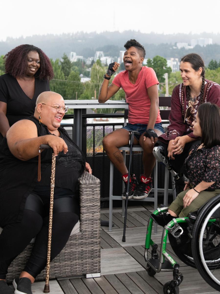 Five disabled people of color with canes, prosthetic legs, and a wheelchair sit on a rooftop deck, laughing and sharing stories. Greenery and city high-rises are visible in the background. Credit of the image to Disabled And Here: https://affecttheverb.com/disabledandhere/