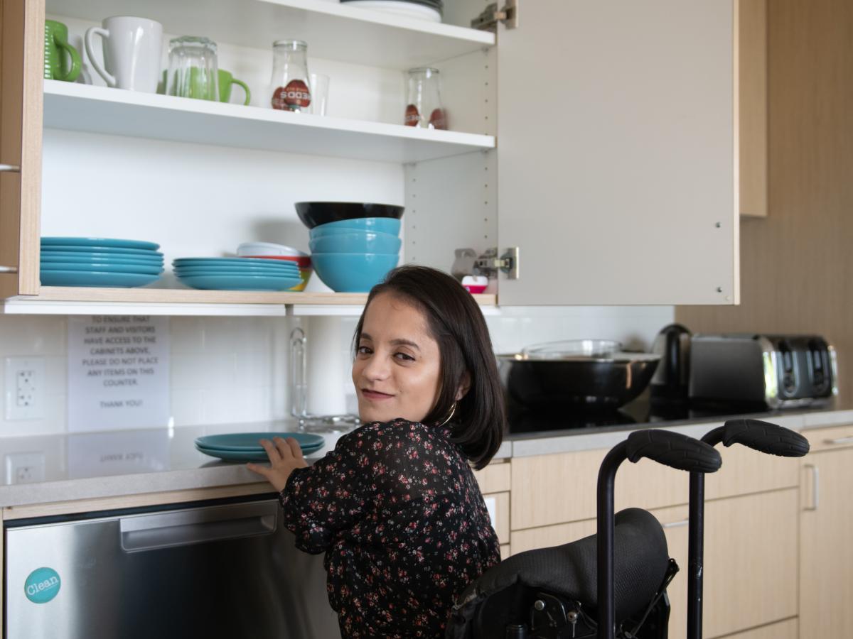 A South Asian person sitting in her wheelchair glances at the camera while grabbing dishes from a lowered height-adjustable kitchen shelf.